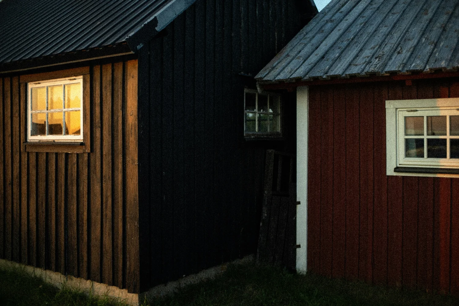 Photo of two wooden houses with sloped roofs, one dark brown and one red, at dusk. The brown house has a window with a yellowish glow, the red house has a window with a white frame, and a small window with a dark interior is visible between them.