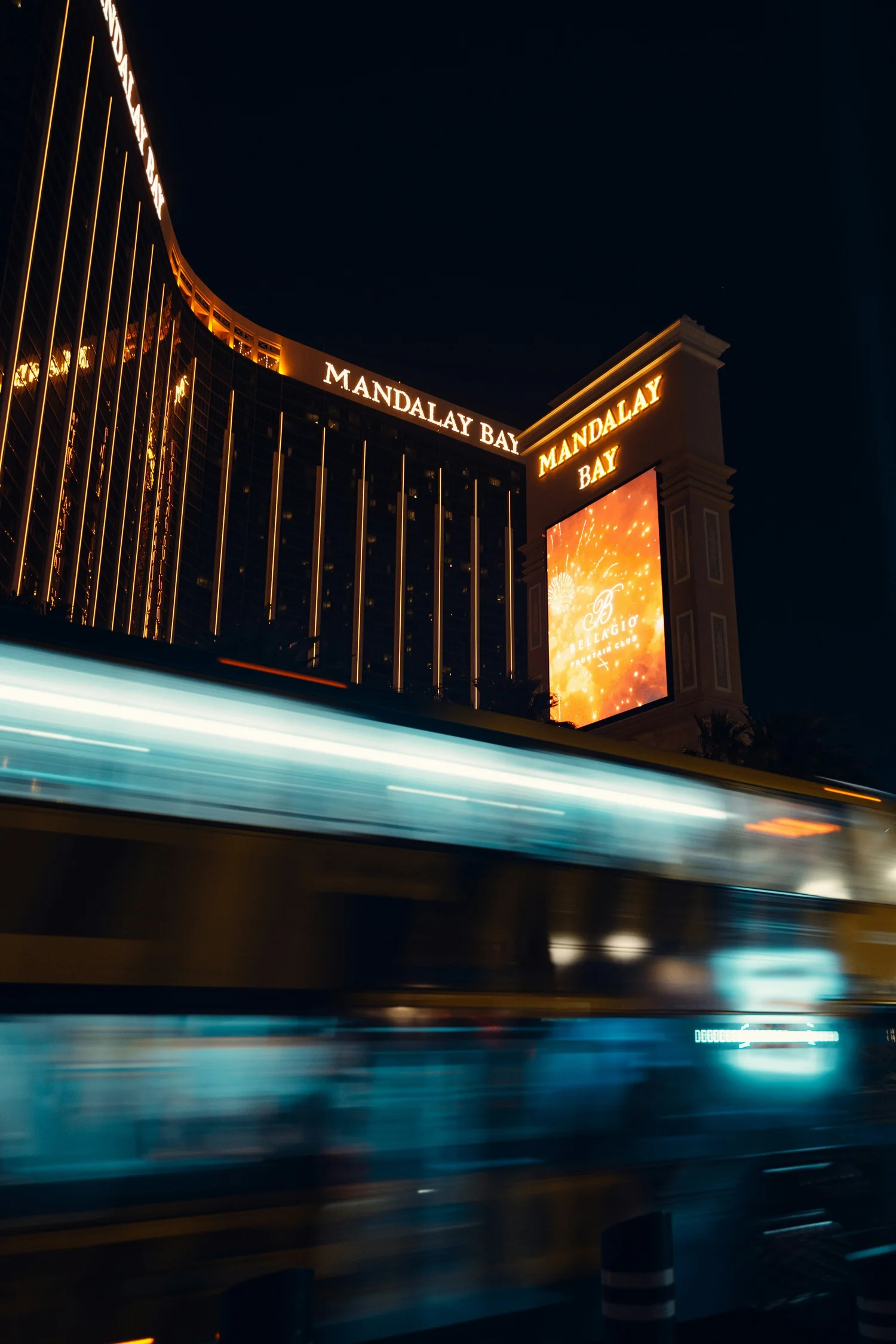 Nighttime view of the Mandalay Bay hotel and casino in Las Vegas, with illuminated signs and blurred movement of a passing bus.