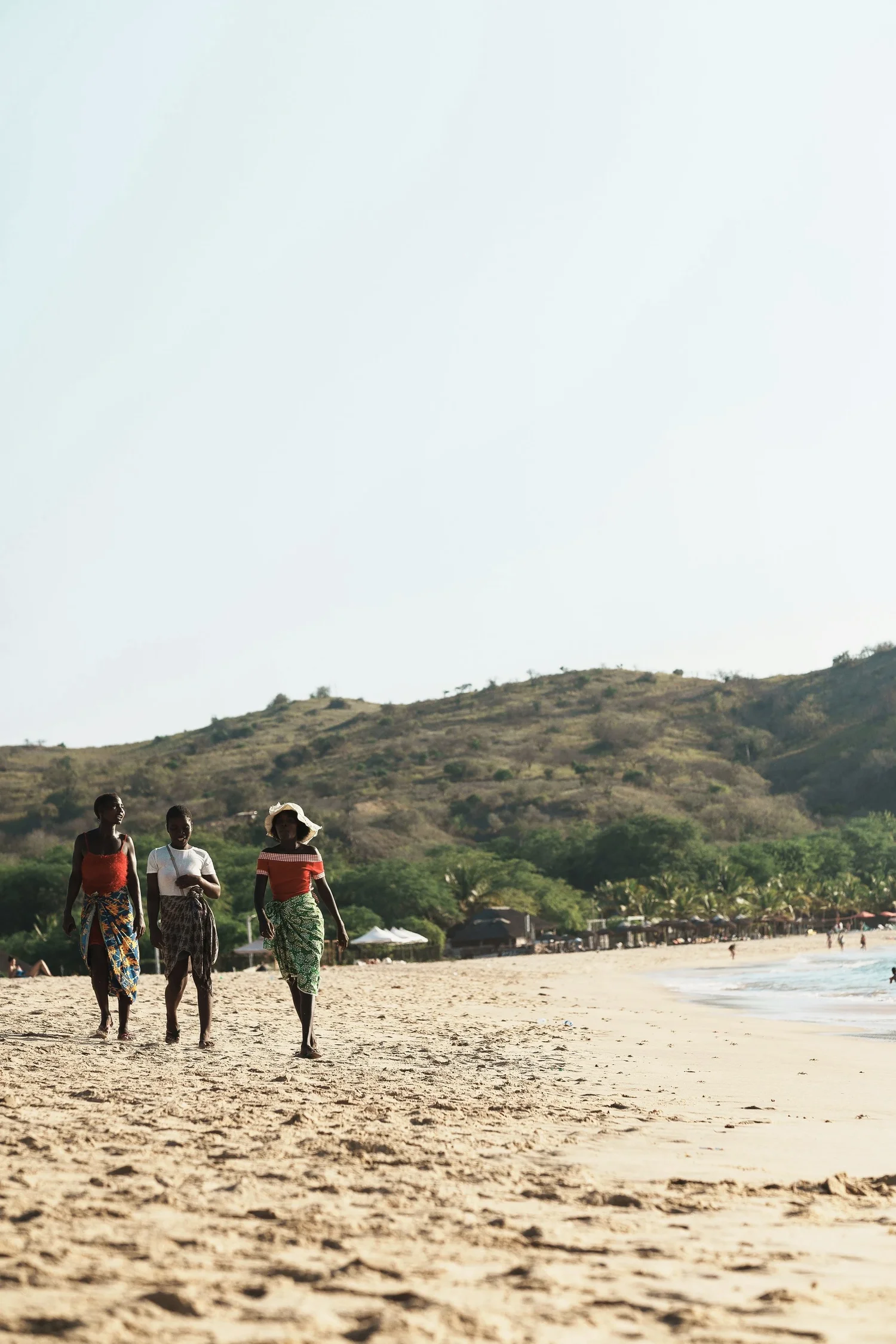 Three women walking along a sandy beach with a hill and trees in the background.