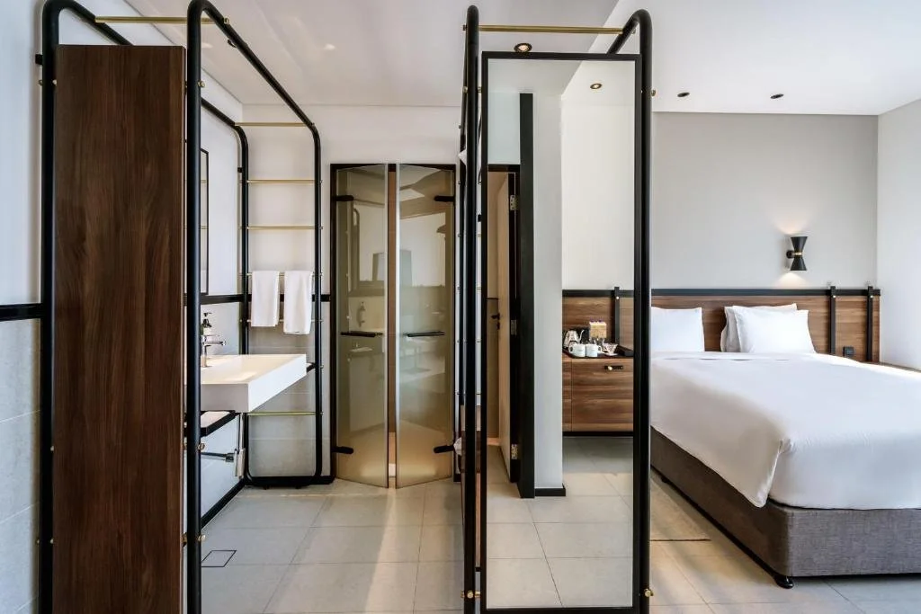 Modern hotel room with a bed, nightstand, and wall sconce, separated from the bathroom area by a black room divider with gold accents, featuring a sink and shower.