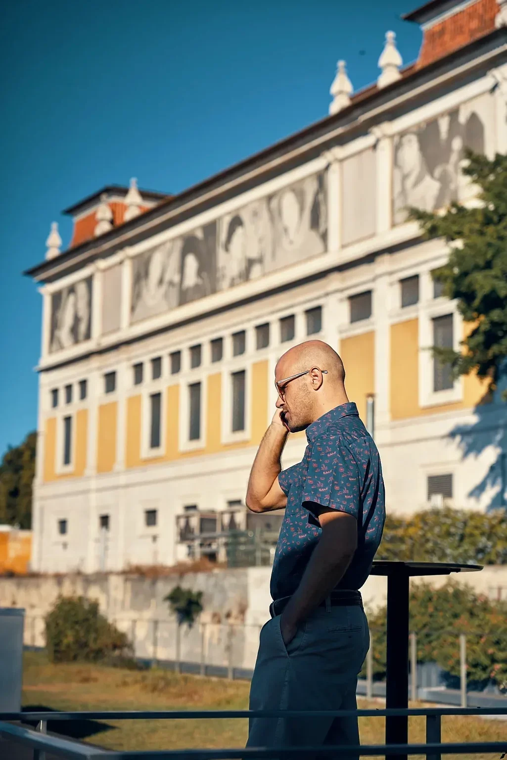 A man with a shaved head and glasses, wearing a patterned short-sleeve shirt, stands outside talking on a cell phone near a historic yellow and white building.
