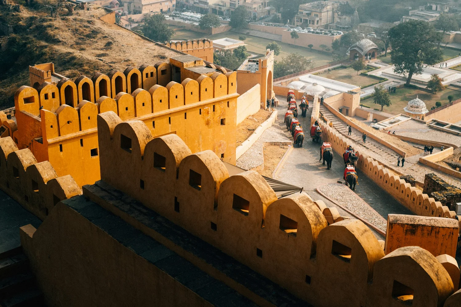 View of Amber Fort in Jaipur, India, showing orange and yellow sandstone walls, a procession of elephants leading up the entrance, and the surrounding courtyard and gardens in the background.