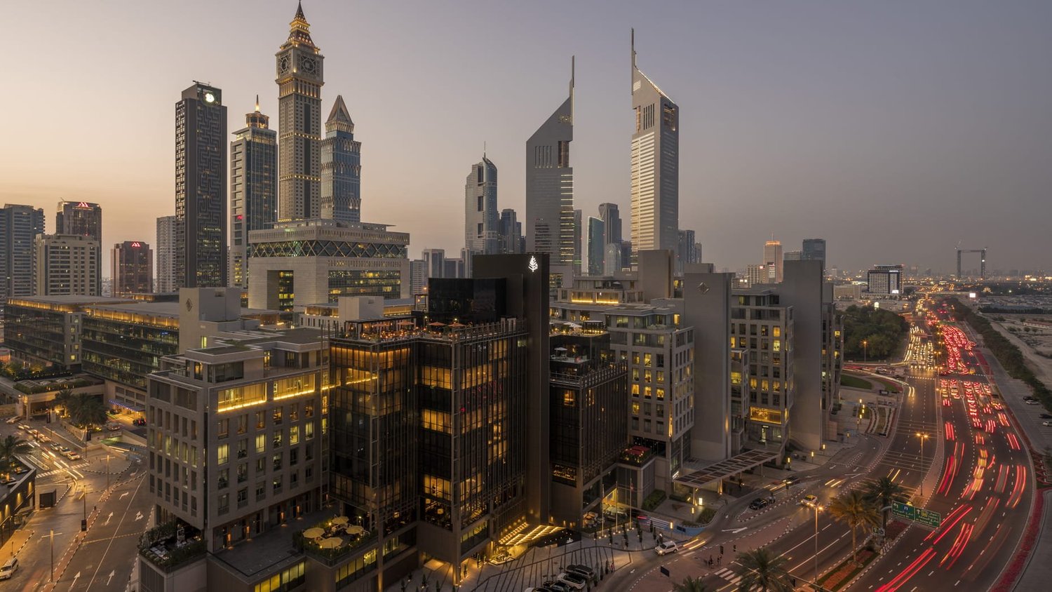 City skyline at dusk with high-rise buildings, some with illuminated windows and signs, busy street with car lights, and modern architecture in Dubai.