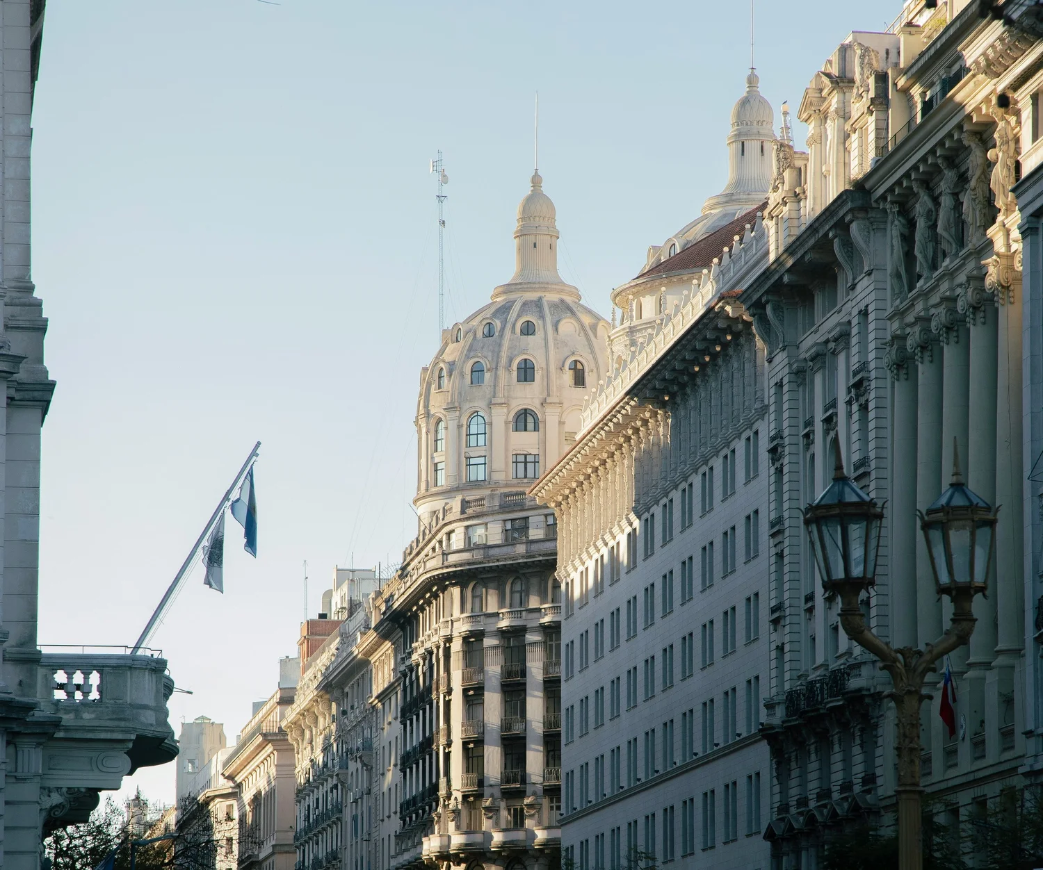 City street with historic buildings and a dome in the background, flags on the left, decorative street lamps on the right, early morning or late afternoon sunlight.