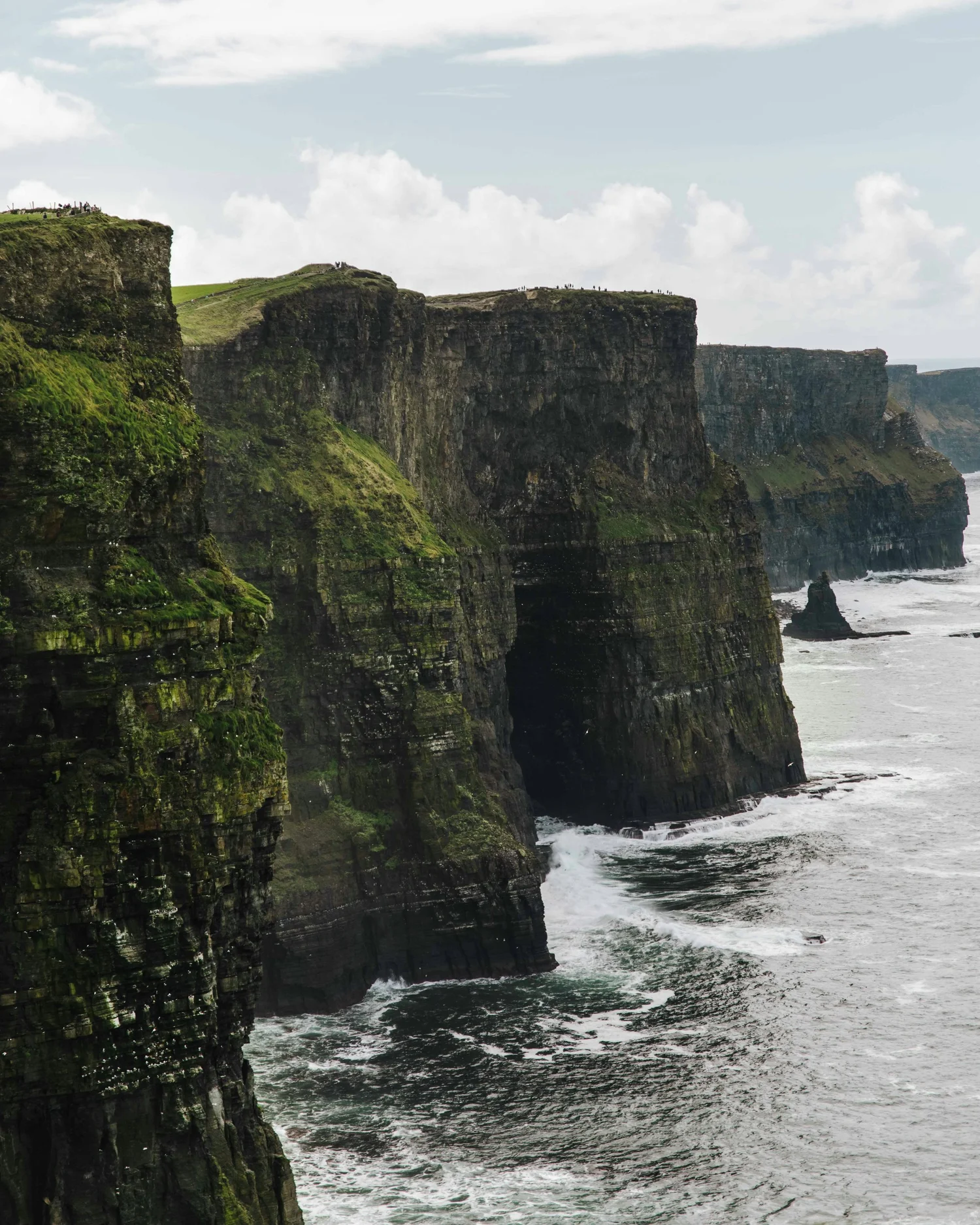 Cliffs along the coast with lush green vegetation, crashing waves below, and a partly cloudy sky.