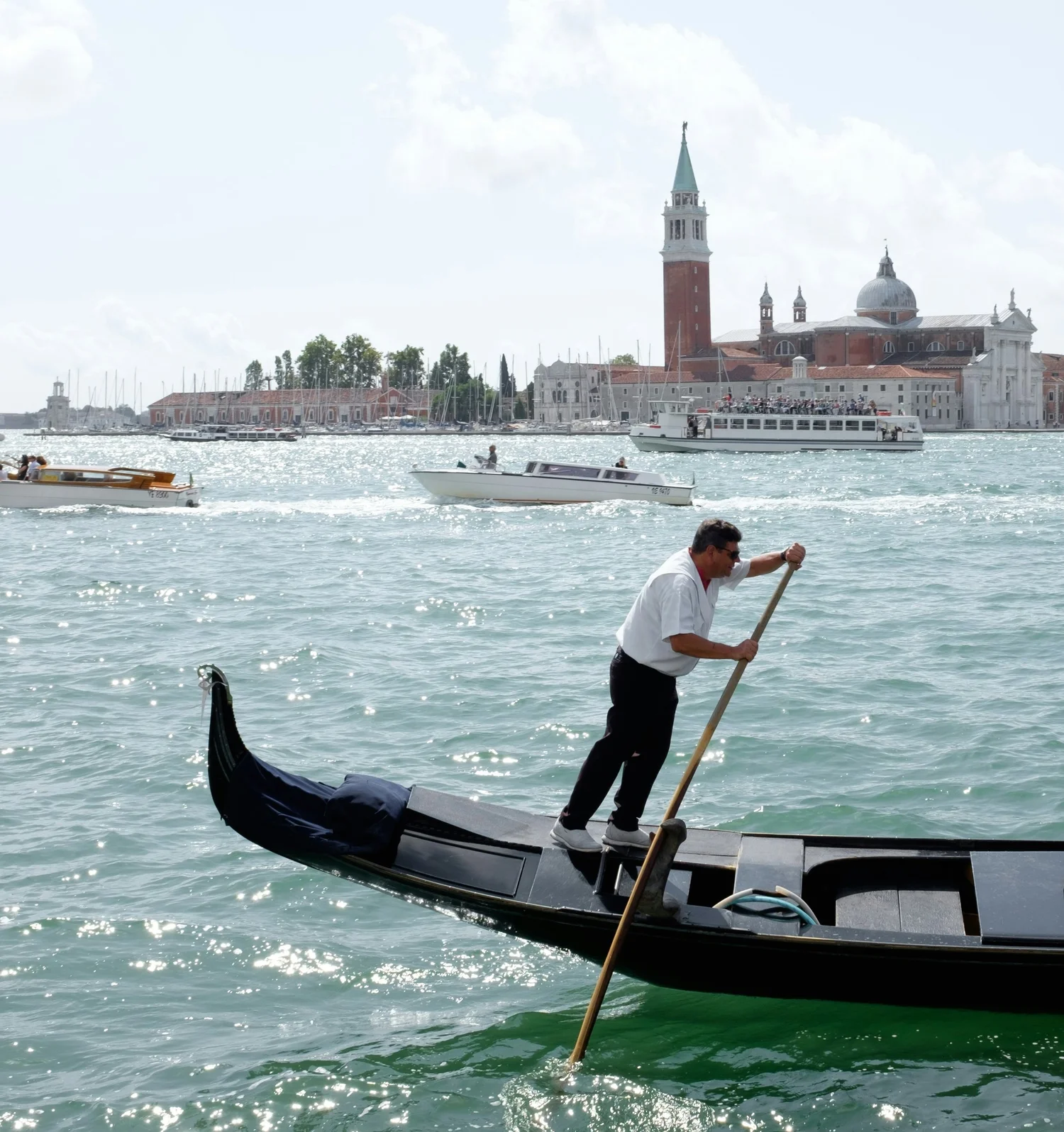 Man in white shirt standing on a gondola boat in Venice, Italy, with water, buildings, and boats in the background.