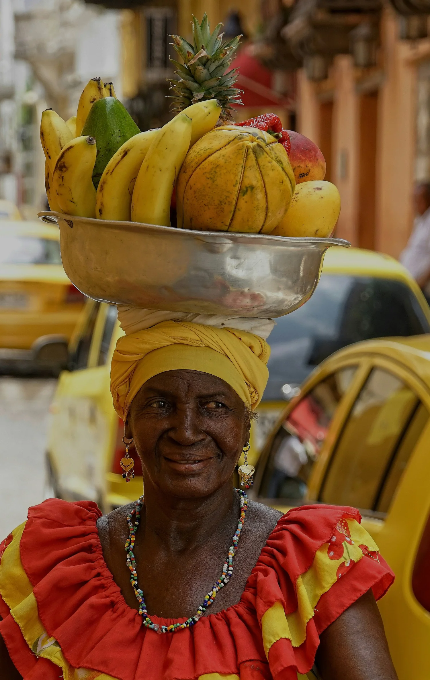 A woman with dark skin wearing a yellow headscarf and a red and yellow dress, balancing a large metal tray of assorted tropical fruits, including bananas, pineapple, and a pumpkin, on her head, on a street with yellow vehicles in the background.