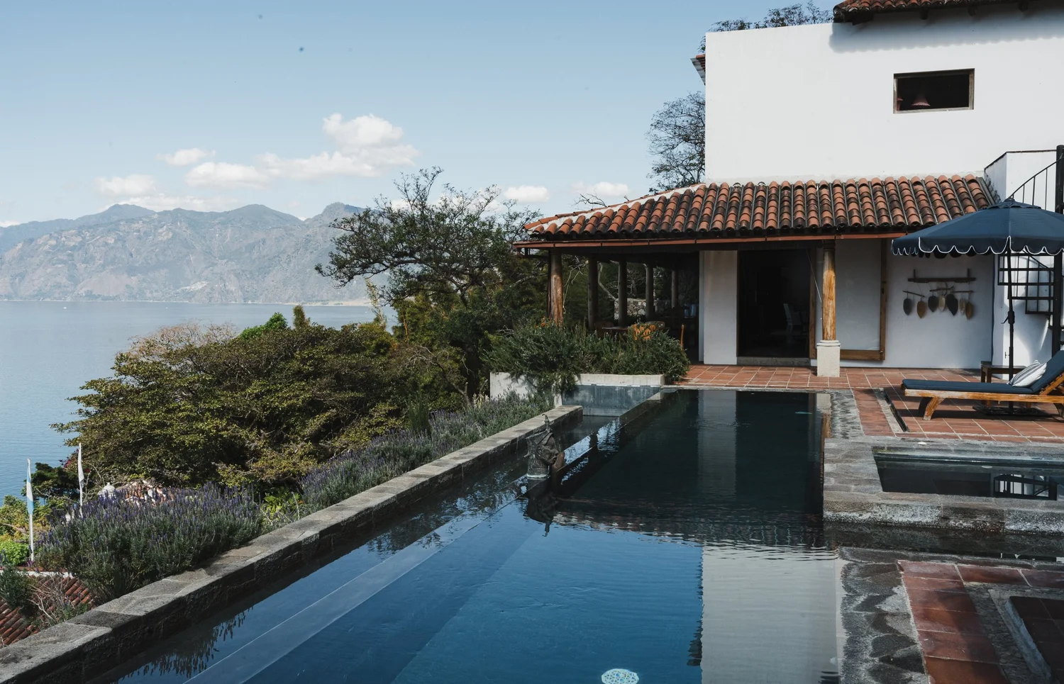 Luxury backyard with swimming pool, patio furniture, umbrella, view of lake and mountains in distance, white house with tiled roof, trees and shrubbery