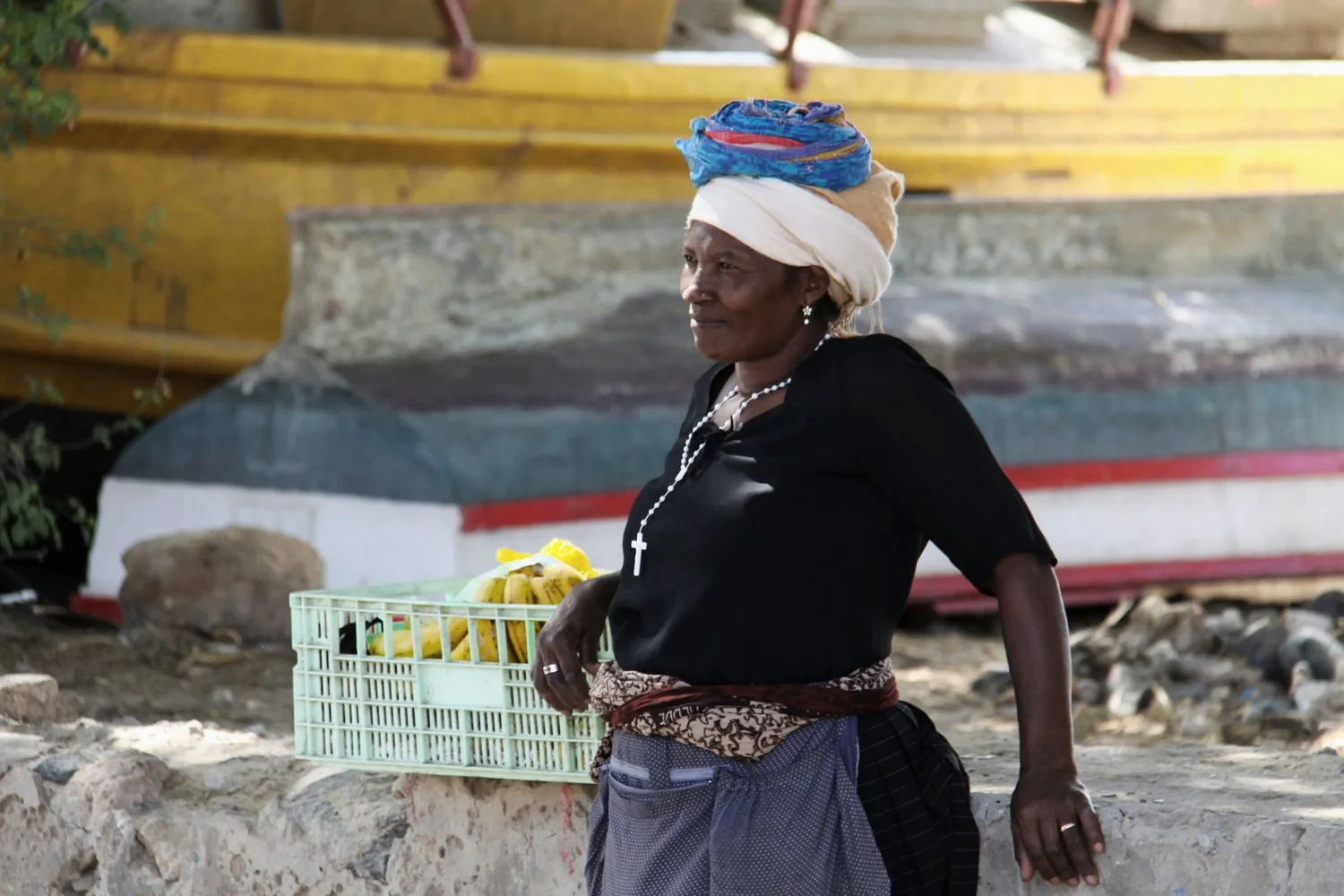 A woman wearing a headscarf and jewelry stands outdoors with a basket of bananas, in front of a boat on a rocky ground.