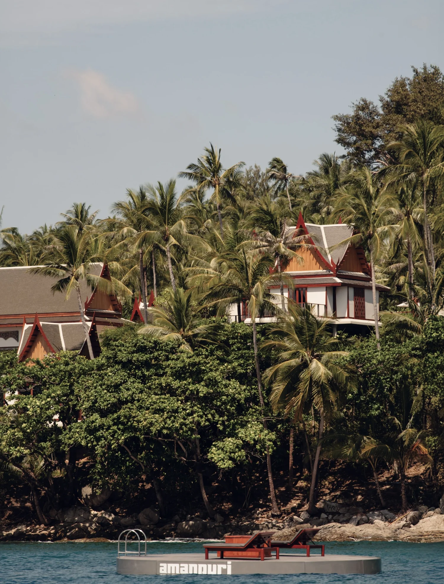 A tropical hillside resort with traditional styled houses situated among palm trees, overlooking water with a floating platform and lounge chairs labeled 'Amanyara' in the foreground.