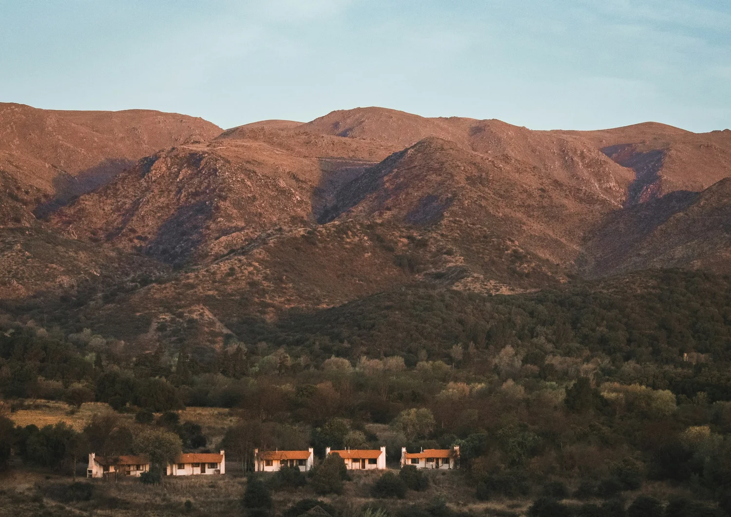 A row of small houses with red roofs and white walls at the base of brown, rolling hills.