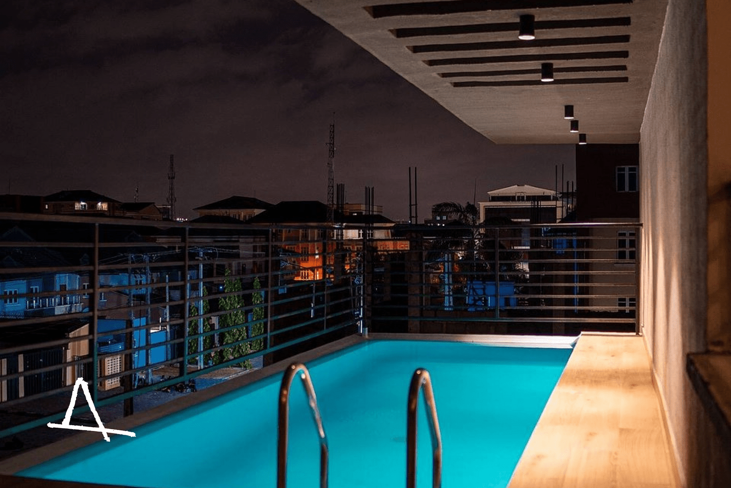 Nighttime view from a balcony with a small illuminated swimming pool, overlooking a city with lit buildings and dark cloudy sky. The accommodation of Lagos Sabbatical, a monthly stay package by Hyperlocal World of Travel.