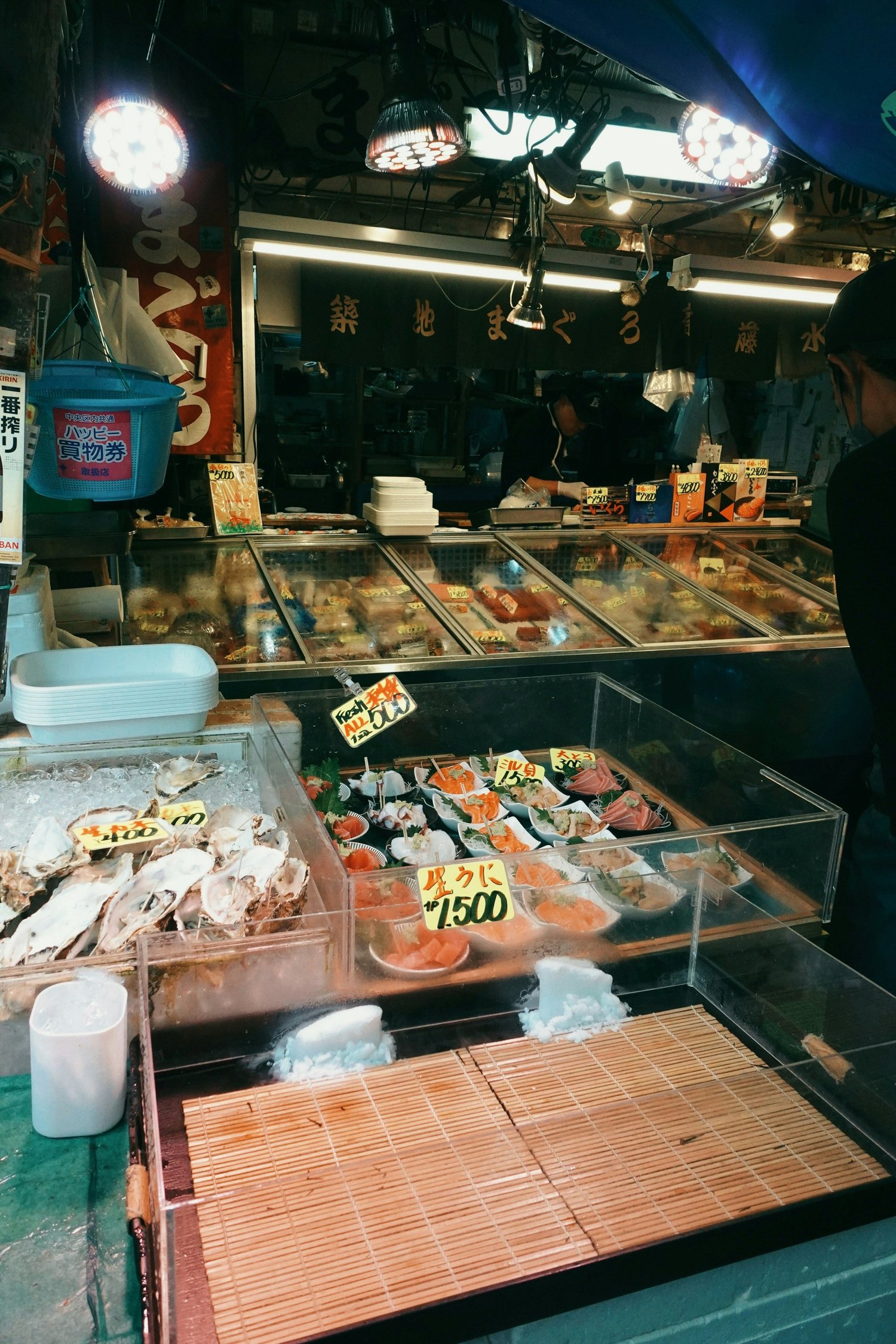 A seafood market stall with oysters, sashimi, and fish laid out on ice and in trays, with handwritten Japanese price tags.