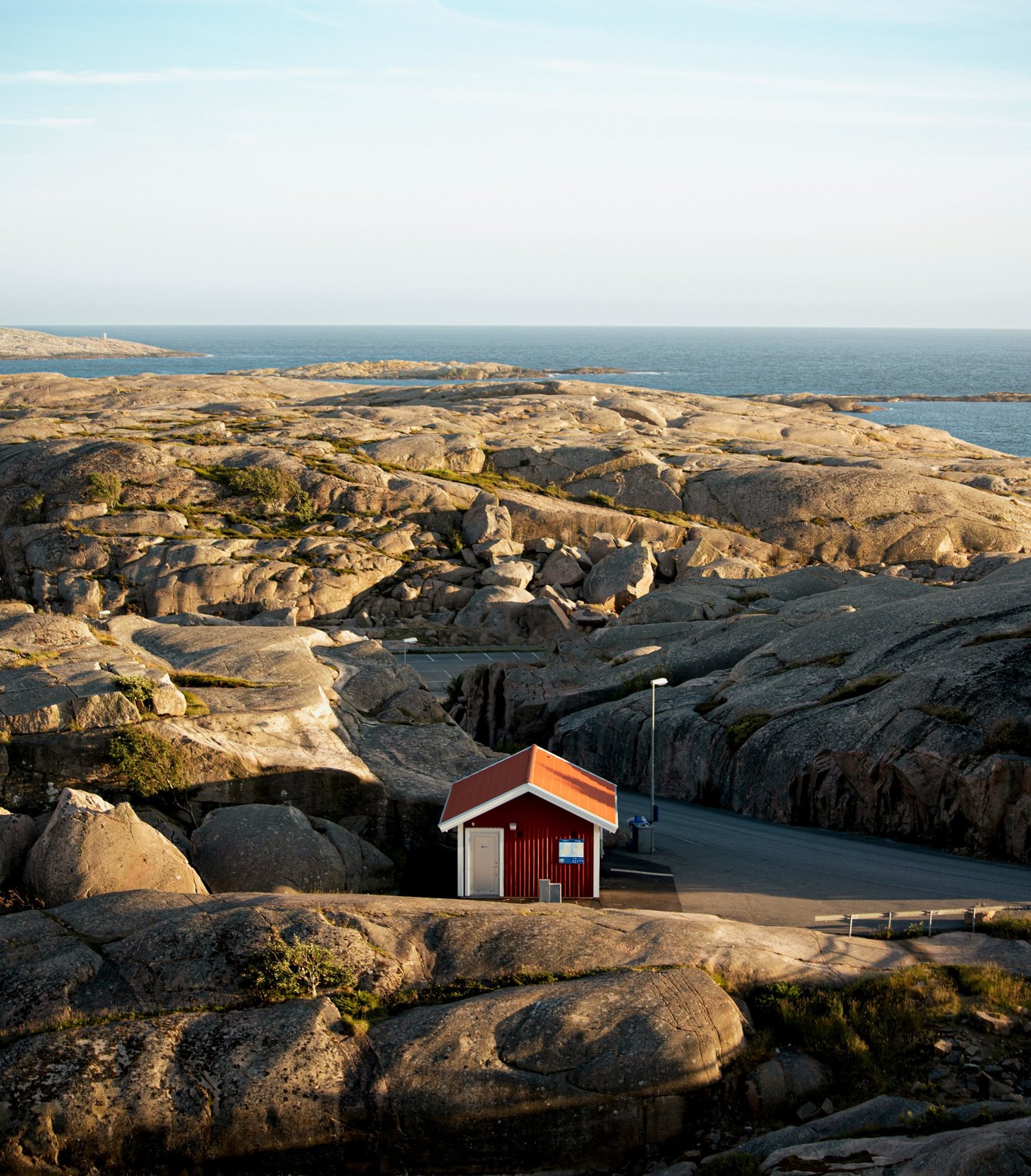 A small red house with a orange roof in a rocky coastal landscape, with the ocean and clear sky in the background.