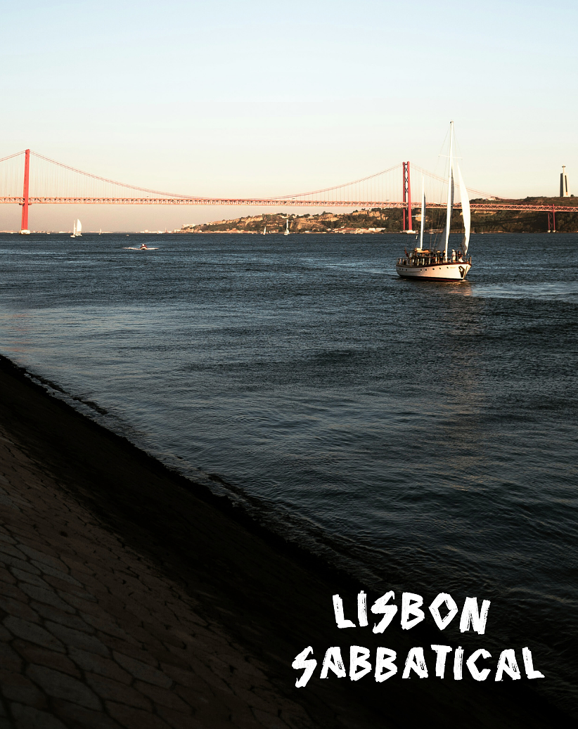 View of the Lisbon waterfront with a sailboat in the water and the 25th April Bridge in the background.