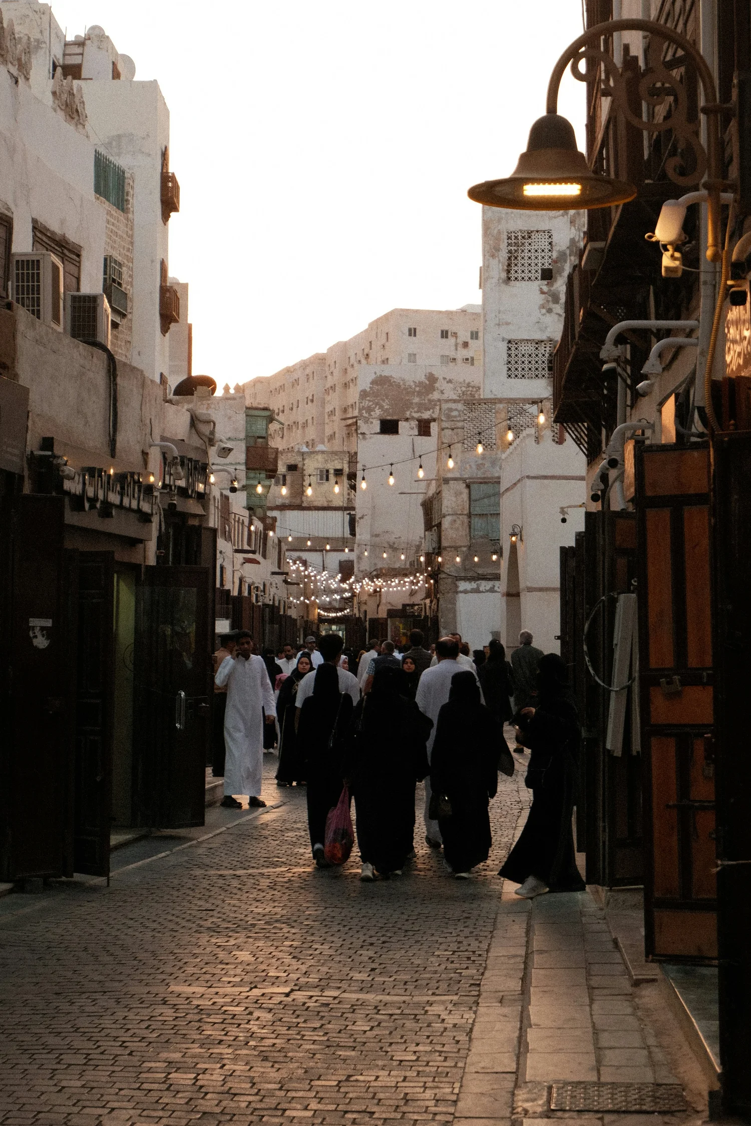 People walking in a narrow street lined with buildings, with string lights hanging across the street at dusk.