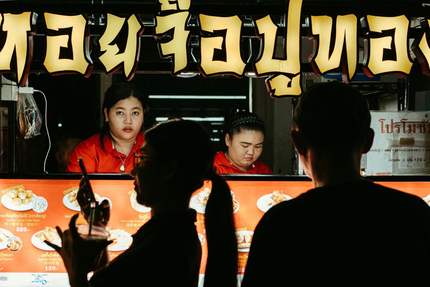 Night market food stall with two women in red uniforms serving customers. Customers are seen in silhouette, one holding a drink with a straw. The stall's menu with images of dishes is visible below the counter. Large illuminated Thai sign is above the stall.