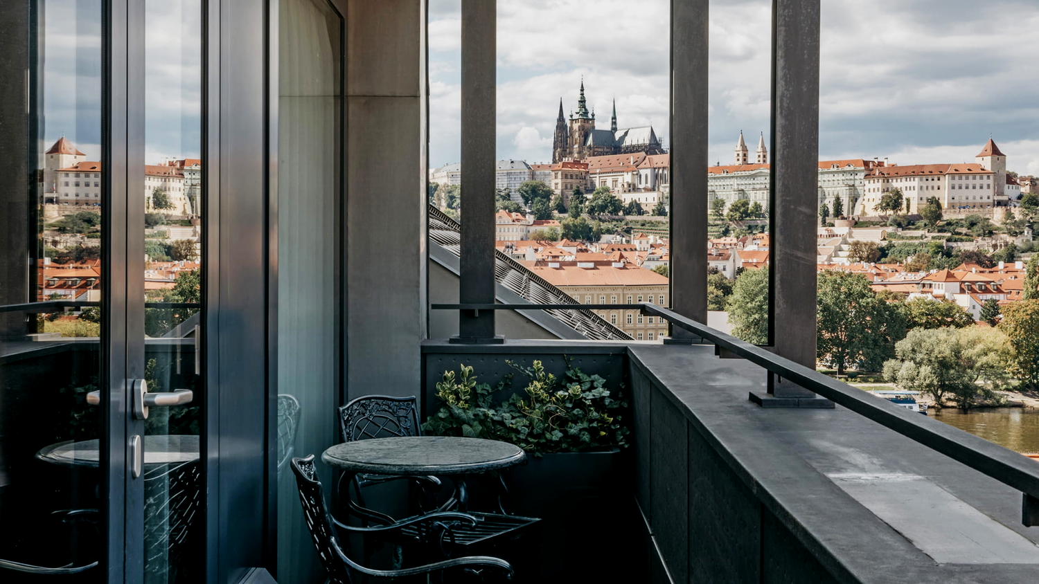 View of a city with historic buildings and a church with tall spires seen from a balcony with black metal railing, small table, and chairs, through large glass windows on an overcast day.