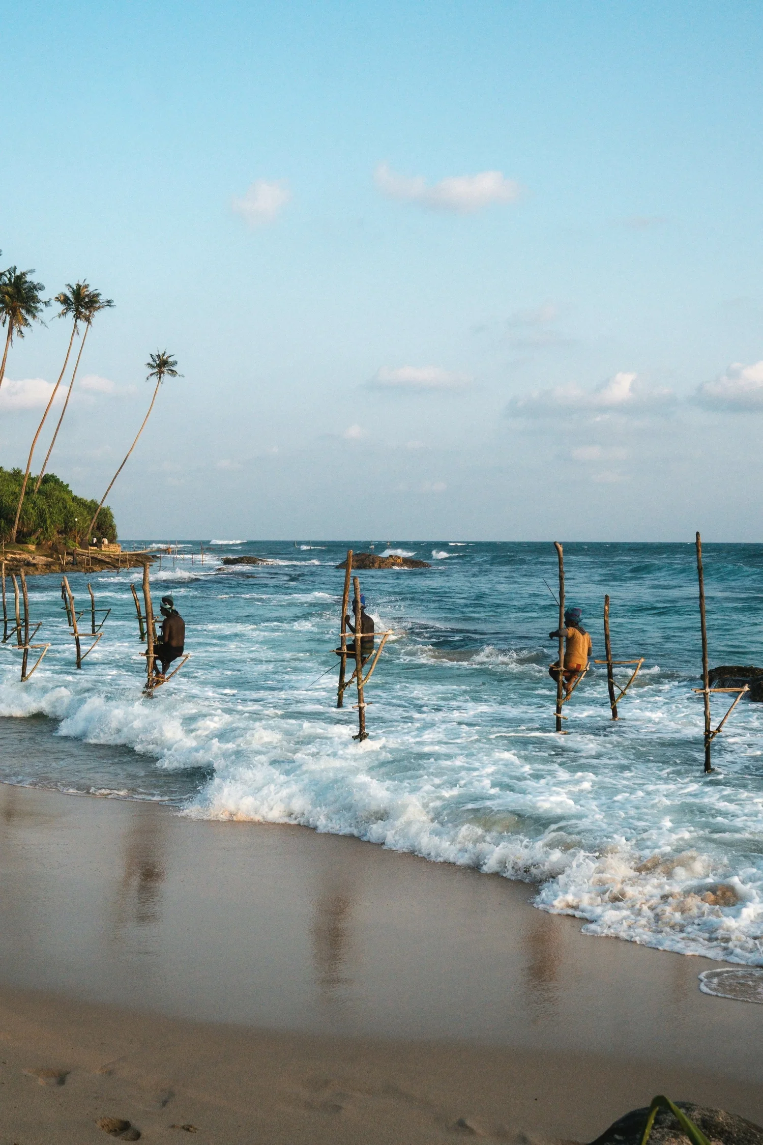 Three people sitting on wooden stilts in the ocean near a sandy beach, with a clear blue sky and palm trees on the left side of the image.