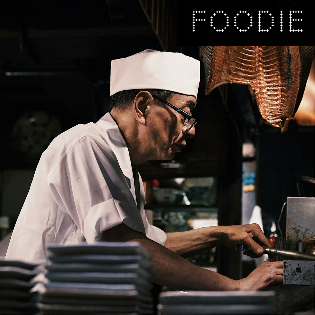 A chef wearing a white hat and coat is preparing food in a kitchen with a dark background, a pig's head hanging overhead, and a sign that reads 'FOODIE' at the top.