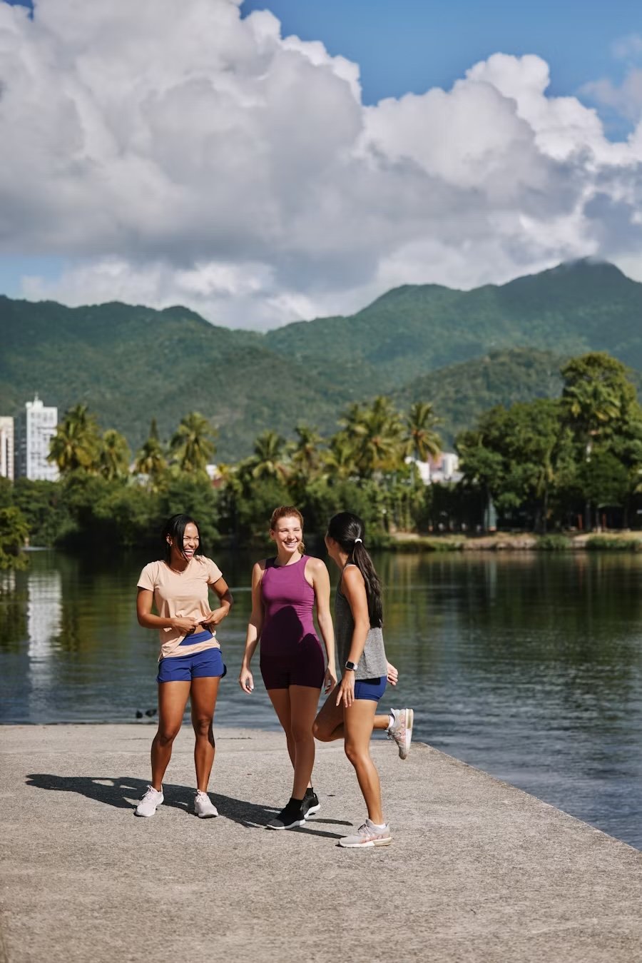 Three women standing and talking near a body of water, with green trees and mountains in the background and partly cloudy sky. Hyperlocal World of Travel.
