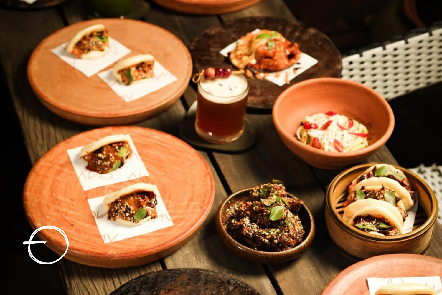 Assorted Asian dishes on wooden and ceramic plates, including tacos, rice bowls, and a drink with cherries on top, spread across a rustic wooden table.