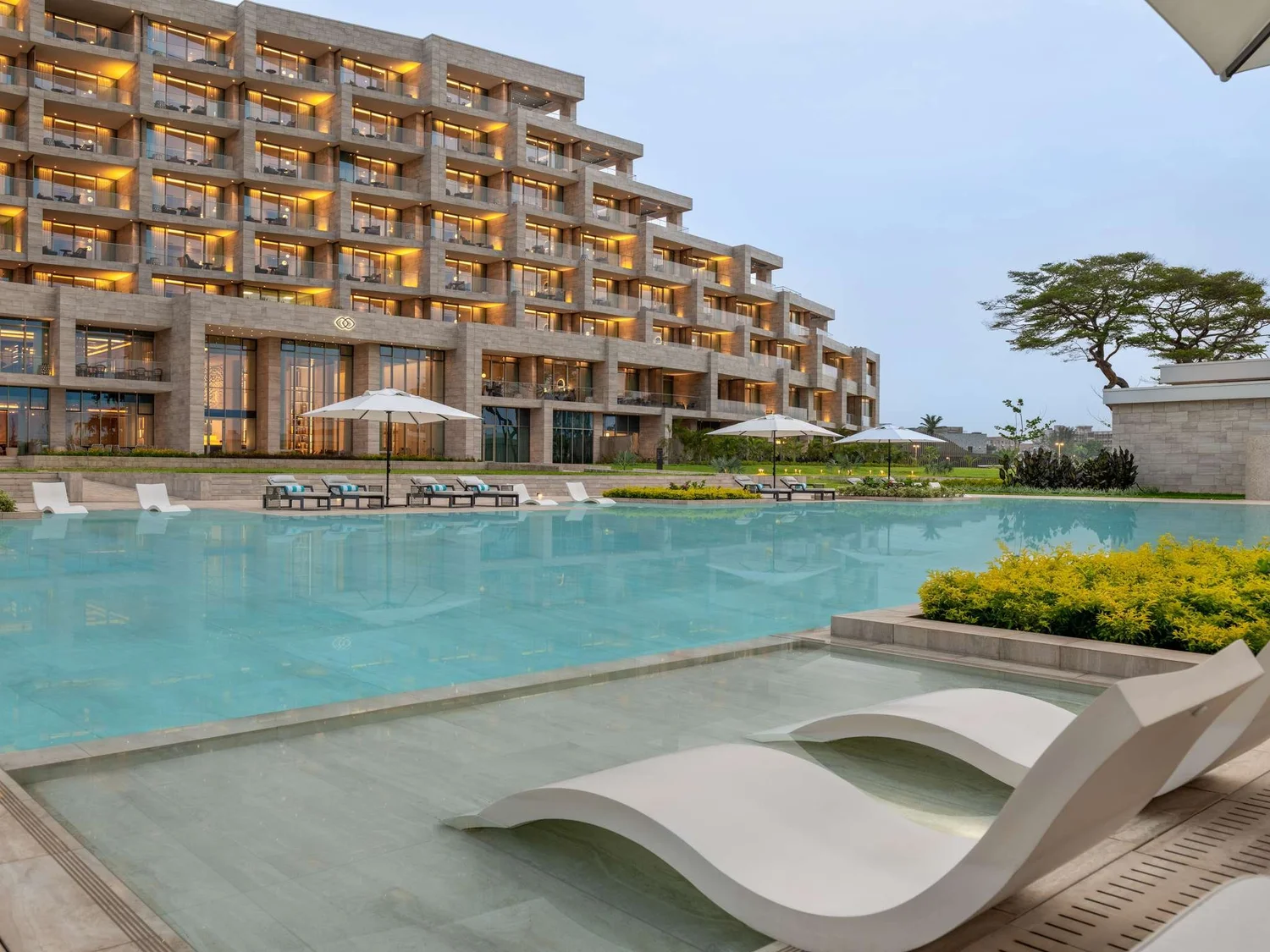 View of a luxurious outdoor swimming pool area with modern lounge chairs and umbrellas, with a multi-story hotel building in the background, illuminated windows, and landscaped greenery surrounding the pool during dusk.