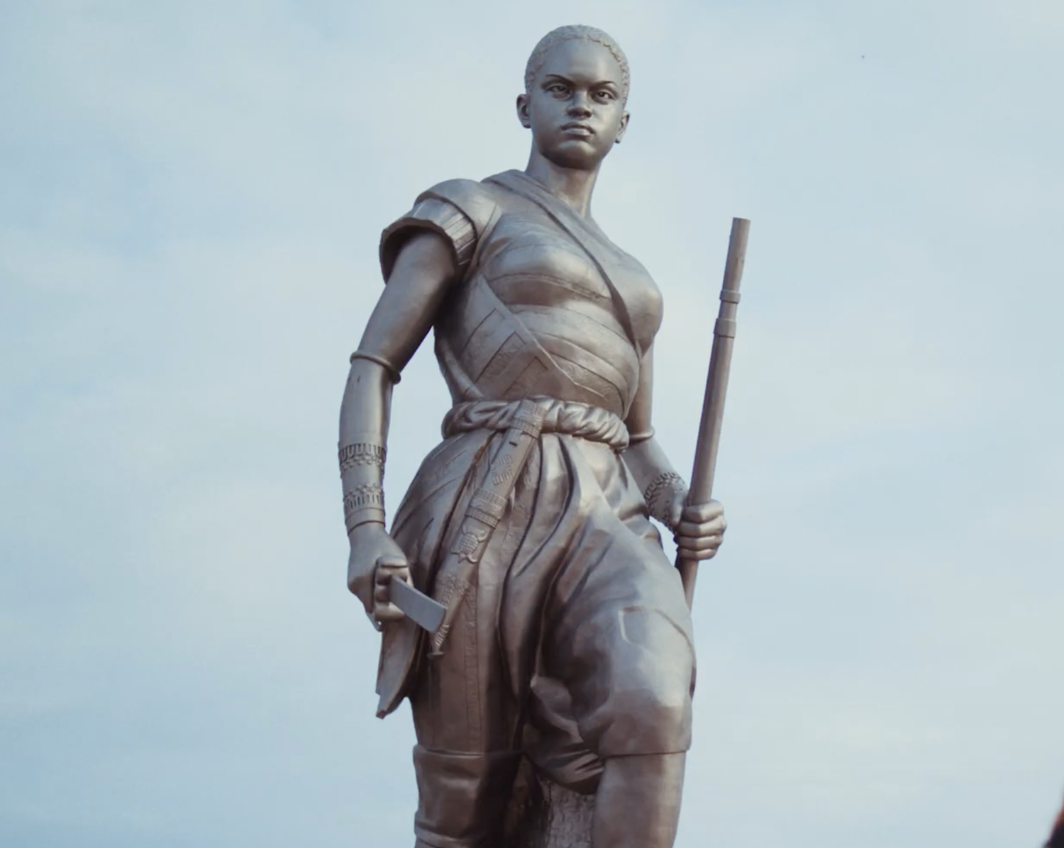 Bronze statue of a female warrior holding a staff and a dagger, dressed in traditional armor, standing against a cloudy sky.