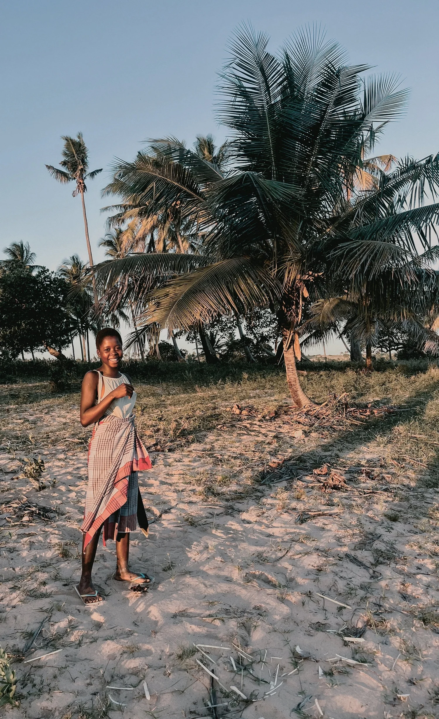 A smiling girl standing on a sandy beach with palm trees in the background during sunset.