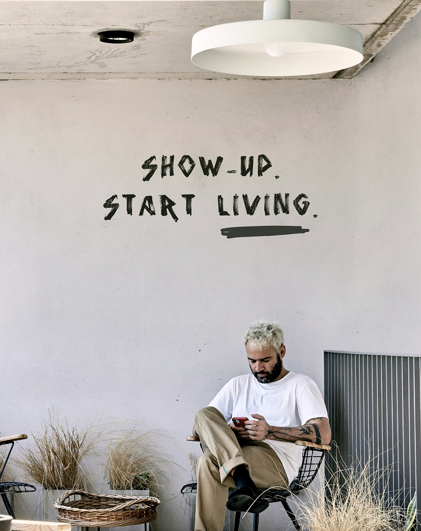 A man with white hair sitting on a black chair, looking at his phone, in a room with a white wall that has the words 'Show Up. Start Living.' written on it, and a ceiling light fixture. Hyperlocal World of Travel. Sabbatical pacakages.