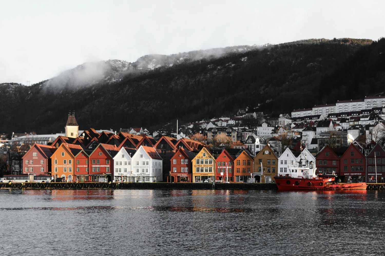 Colorful row of houses along a waterfront with a red boat in the water and mountains in the background.