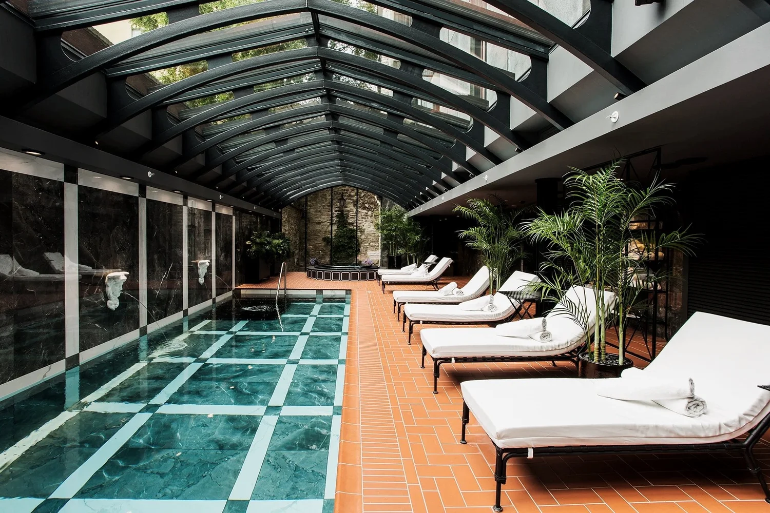 Indoor pool area with lounge chairs, towels, and potted plants under a glass arched ceiling