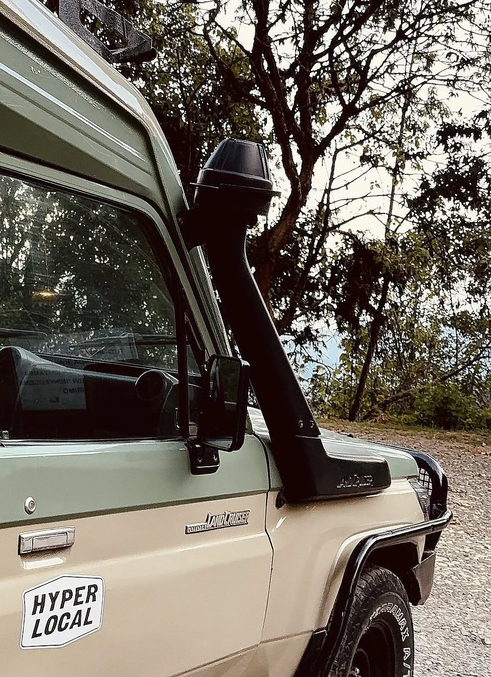 A close-up view of a vintage Toyota Land Cruiser vehicle with a sticker that says 'HYPER LOCAL' on its door, parked outdoors with trees and sky in the background.
