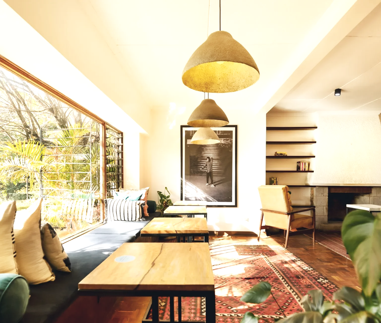 Bright and cozy living room with large window, wooden floor, hanging pendant lamps, a framed black-and-white photograph, a fireplace with shelves, and a mix of cushions and furniture.