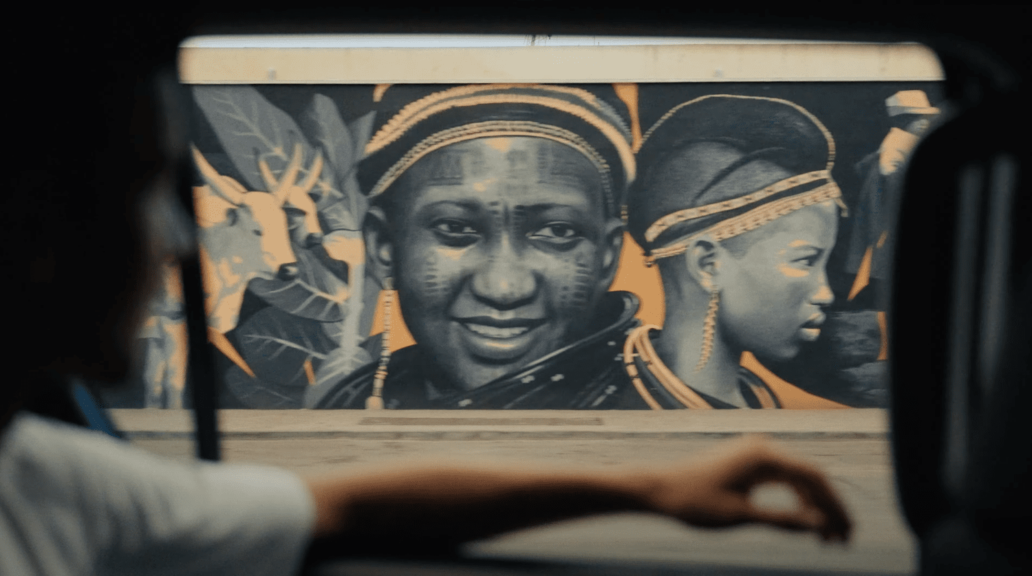 Colorful mural of two African women wearing traditional attire, seen through the window of a vehicle, with a person's arm and head in the foreground.