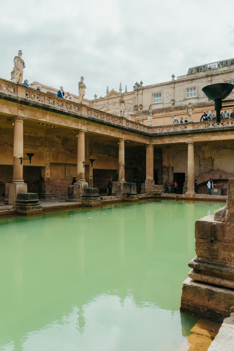 Ancient Roman bath with a rectangular pool filled with greenish water, surrounded by stone columns and arches, with people walking and standing on upper and lower levels.
