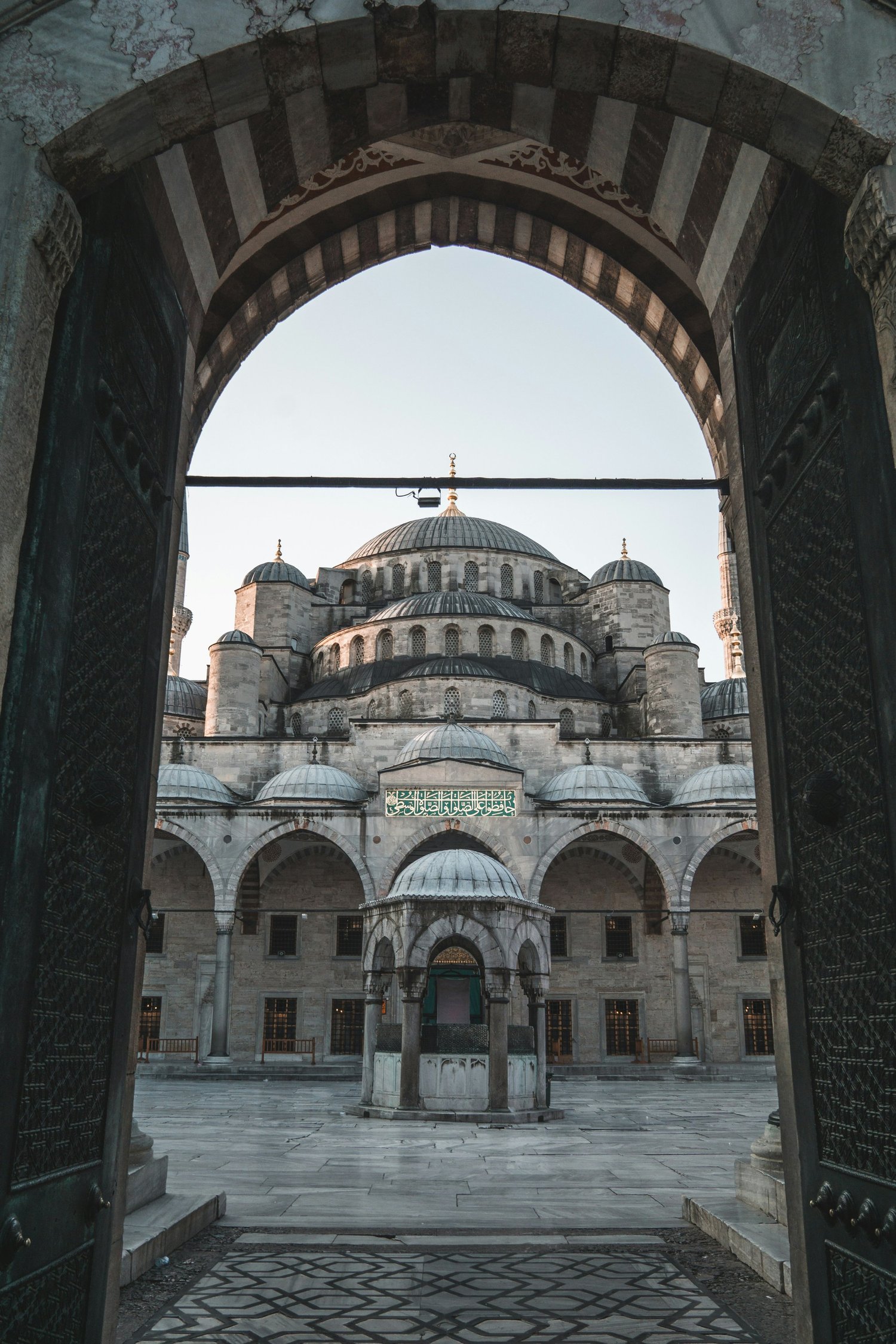 View of a mosque with domes and minarets, seen through an ornate door archway in a courtyard, showcasing Islamic architecture.