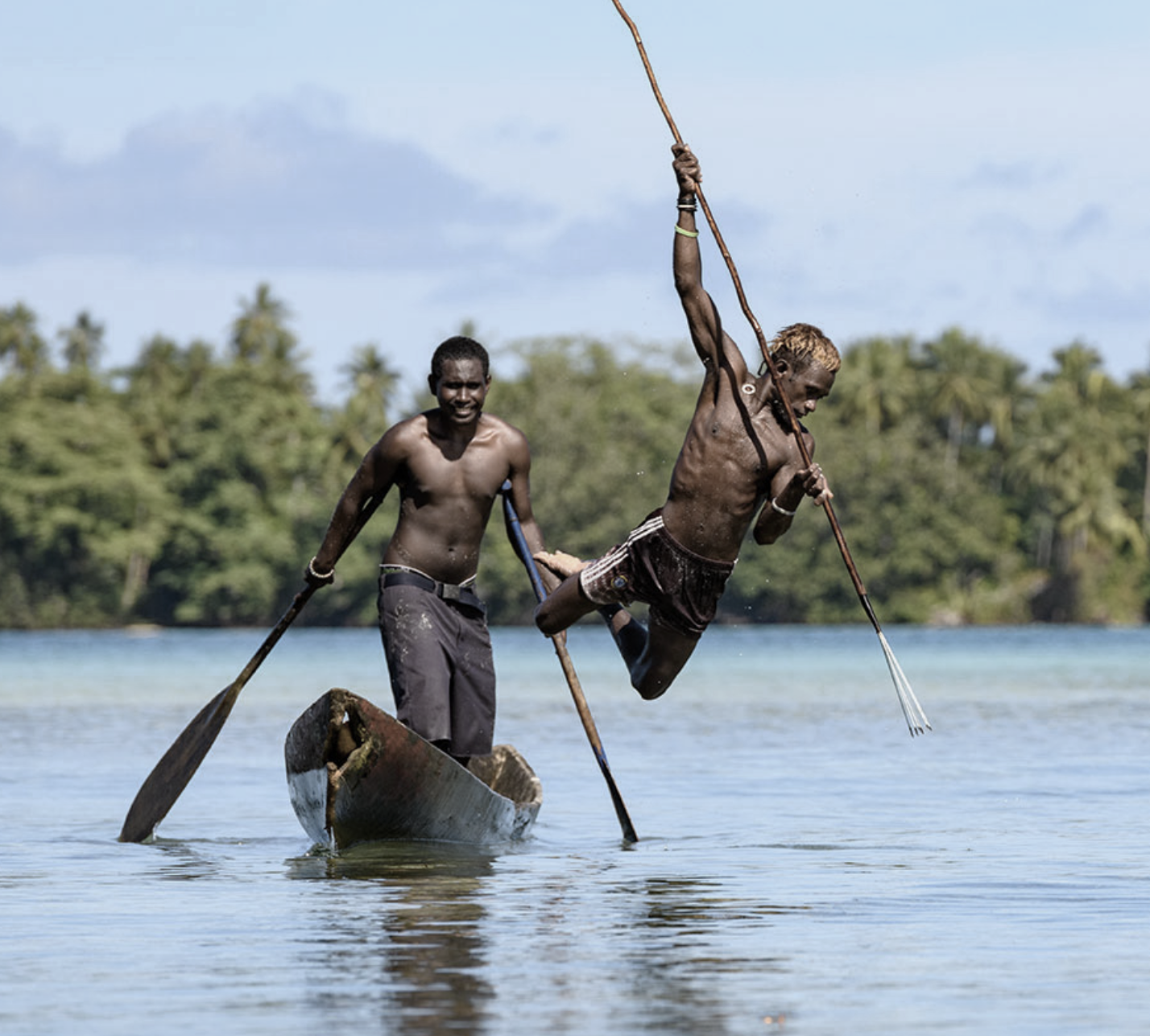 Two young men in a boat on a body of water, one holding a spear and the other paddling, with lush green trees in the background.