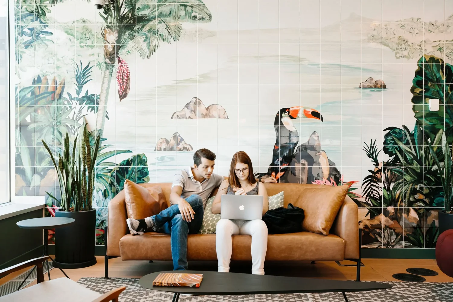 Two young people sitting on a caramel-colored sofa with a large tropical mural featuring a toucan on the wall behind them, surrounded by potted plants and a coffee table with magazines. Hyperlocal World of Travel. WeWork Rio de Janeiro.