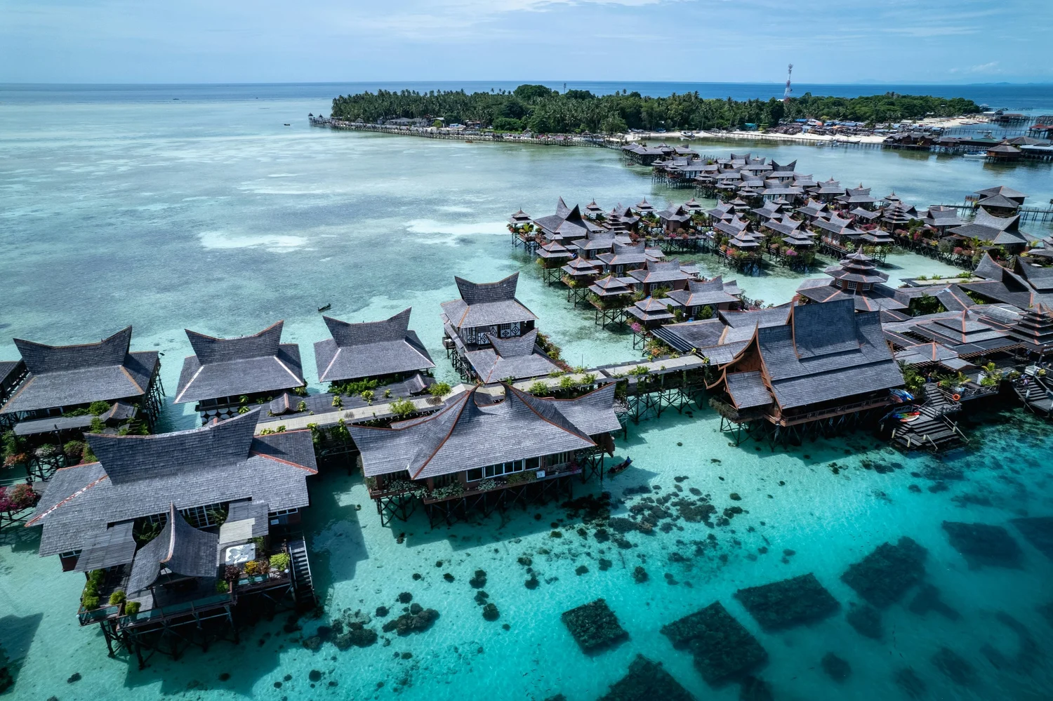 Aerial view of overwater bungalows and villas on a tropical island with clear turquoise water and lush green trees in the background.