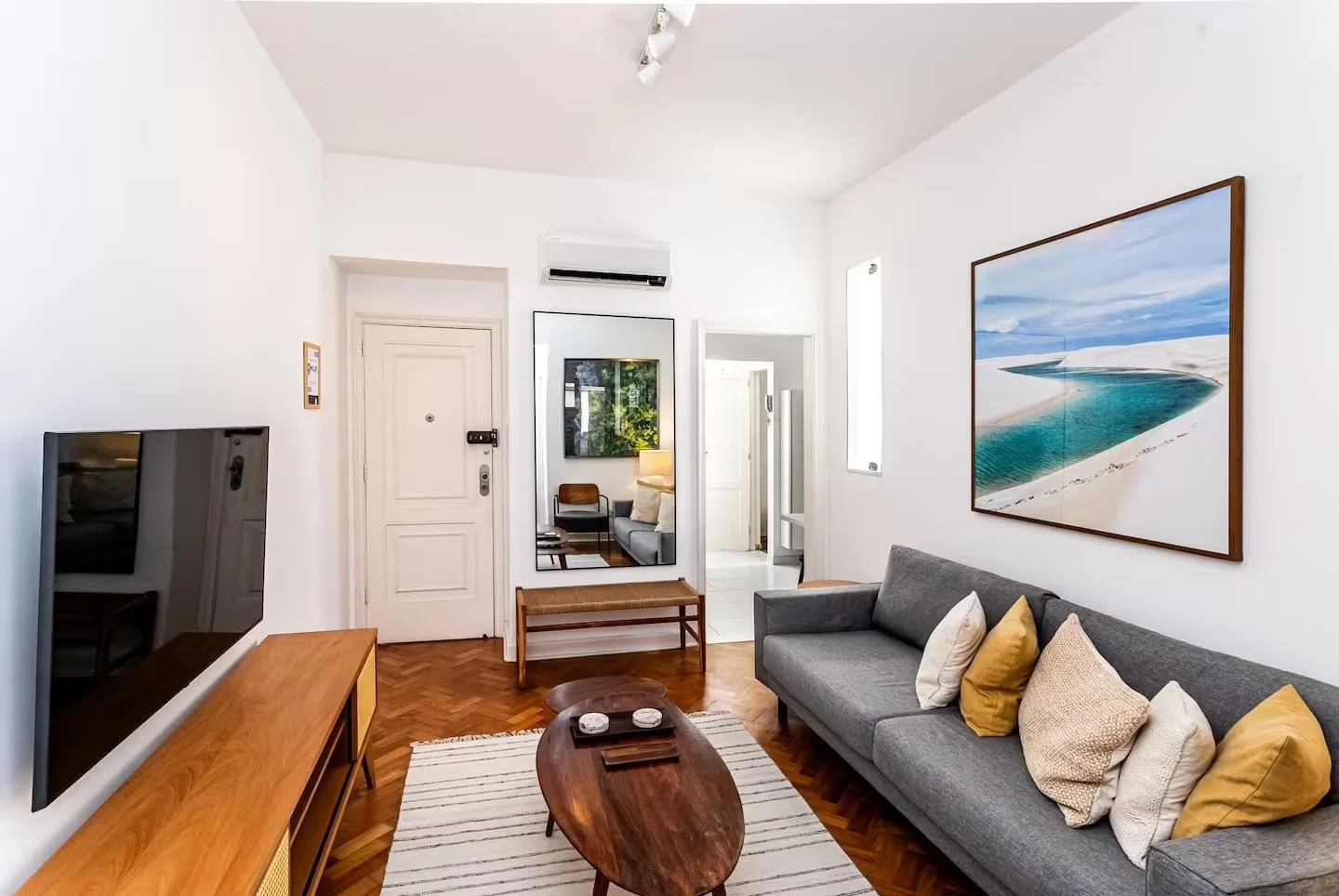 Living room with gray sofa, yellow and white pillows, wooden coffee table, wall-mounted TV, large landscape photo, standing mirror, area rug, white walls, hardwood floor, doorway leading to other rooms, window with natural light.