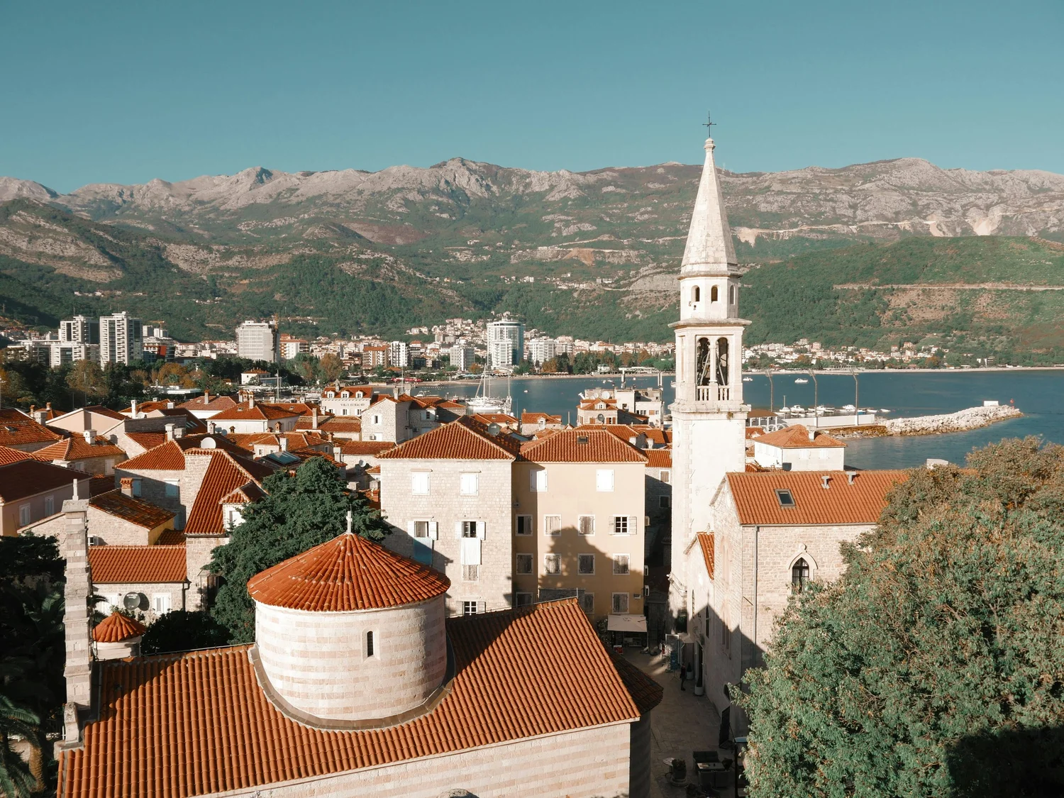 A view of a coastal town with red-roofed buildings, a church with a tall steeple, and mountains in the background. Water and a breakwater are visible along the shoreline.