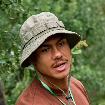 A young man wearing a wide-brimmed, tan hat and a brown shirt, standing outdoors in a green, wooded area.