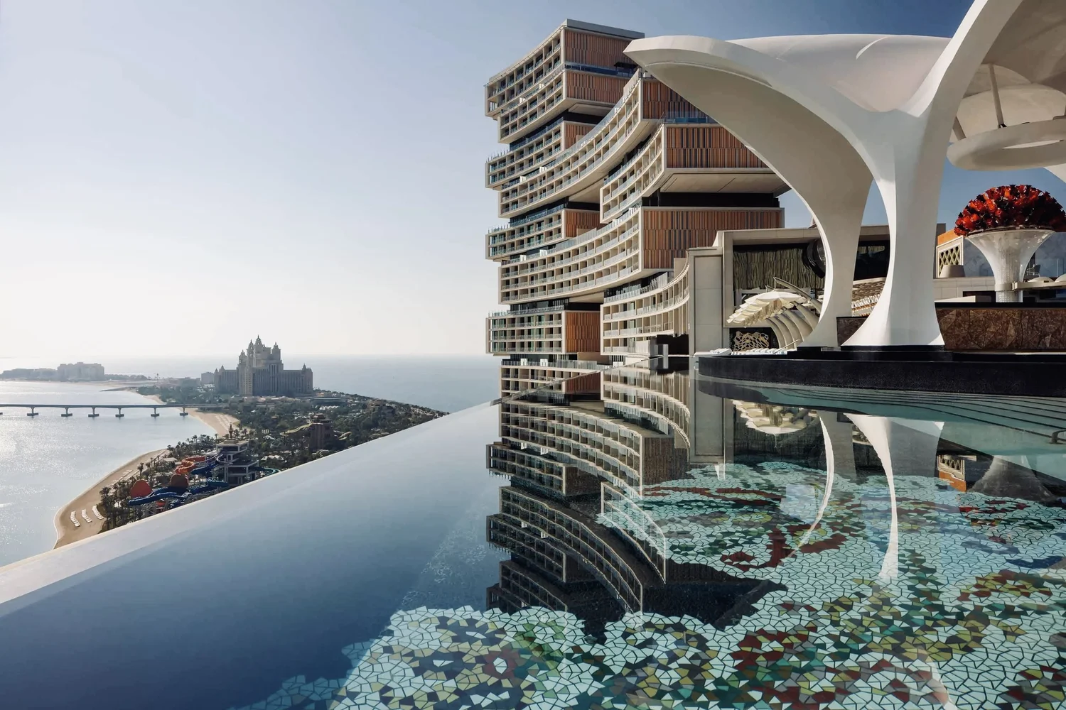 Luxury hotel with a unique, modern architectural design featuring curved balconies overlooking the ocean, with a rooftop infinity pool reflecting the building and sky, and a castle-like structure in the distance near the coast.