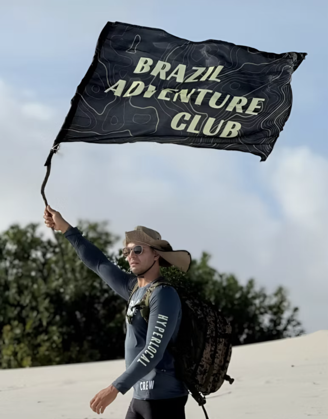 Person holding a black flag that reads 'Brazil Adventure Club' while standing outdoors in a sandy area with trees in the background.