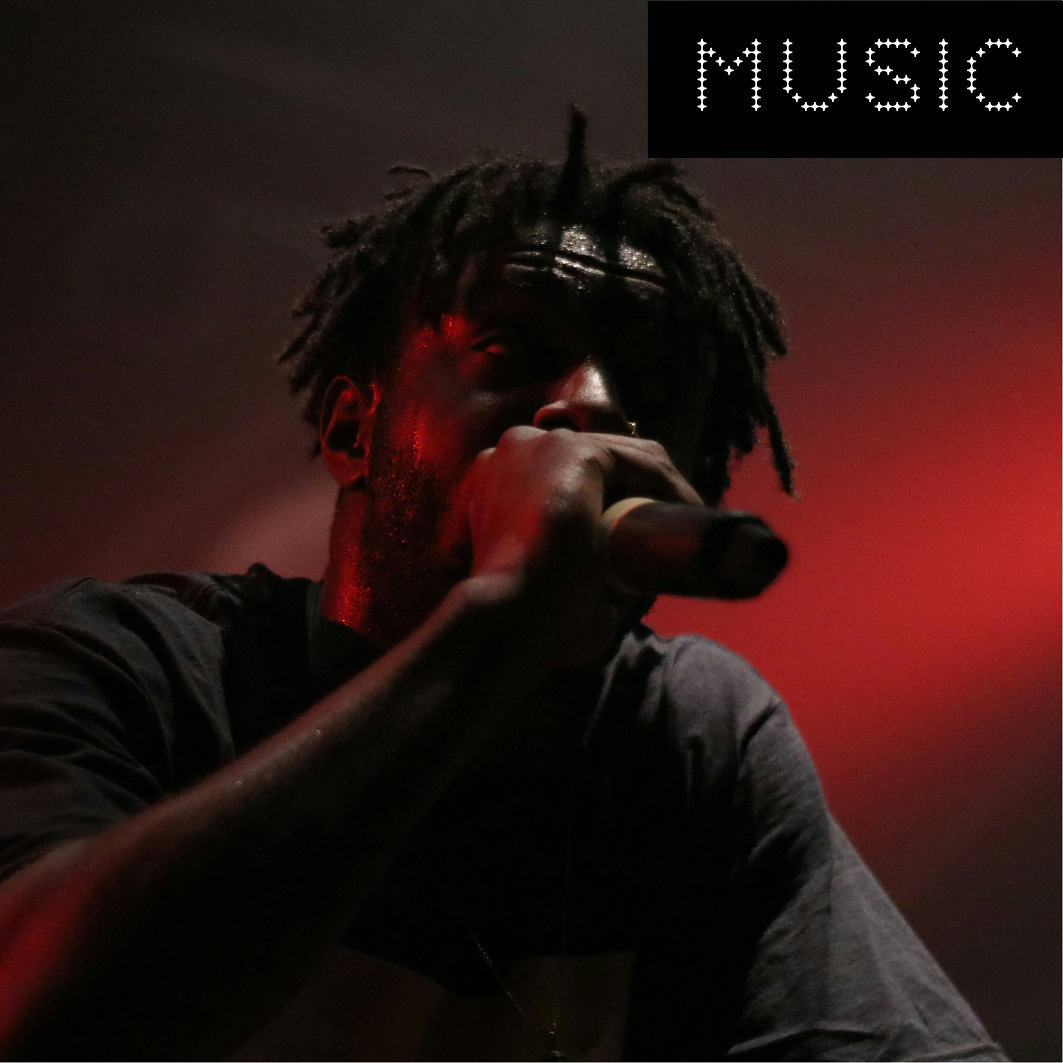 A person with dreadlocks holding a microphone, performing on stage with dark background and red lighting, with the word 'MUSIC' in a stylized font at the top right.