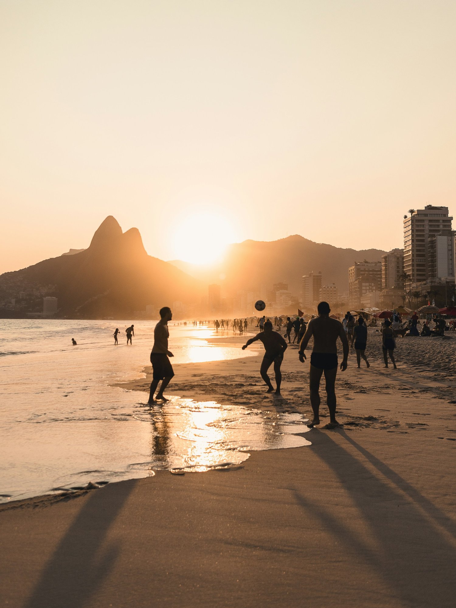 People playing volleyball on a sandy beach during sunset with mountains and city buildings in the background. Hyperlocal World of Travel.