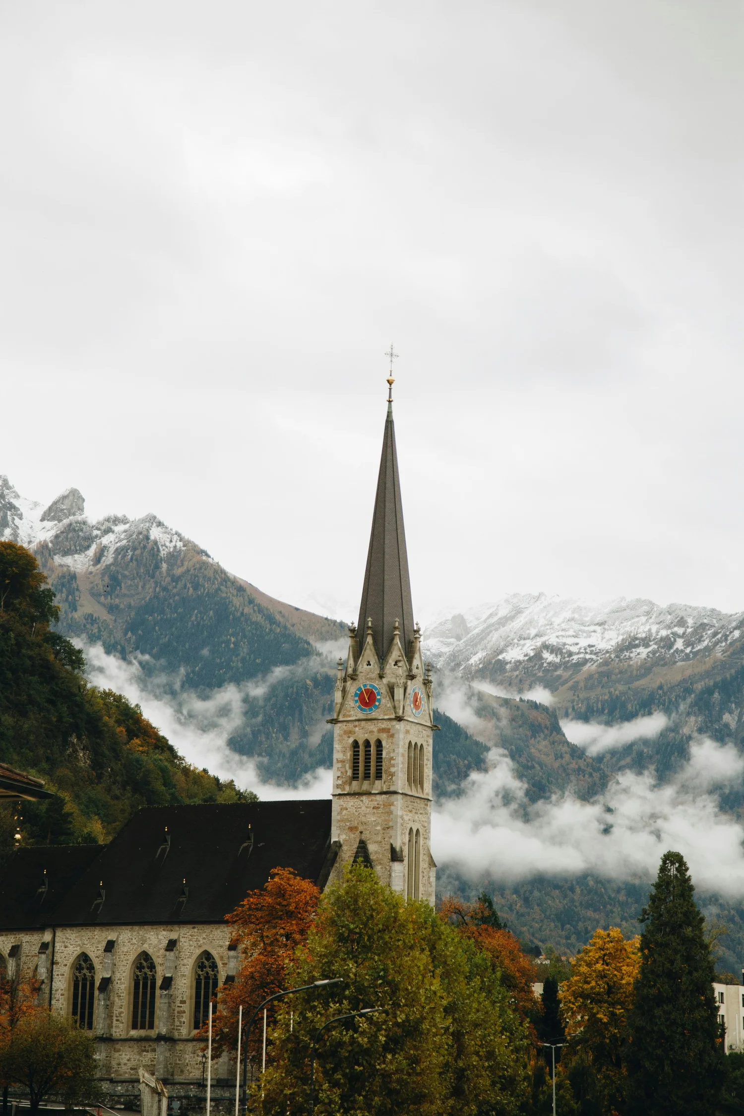 A church with a tall steeple and clock faces, surrounded by fall foliage, mountains, and cloudy sky.