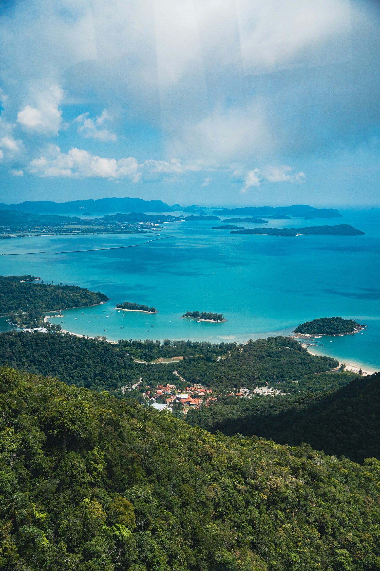 A scenic view of a coastal bay with turquoise waters, surrounded by lush green mountains and islands, under a partly cloudy sky.