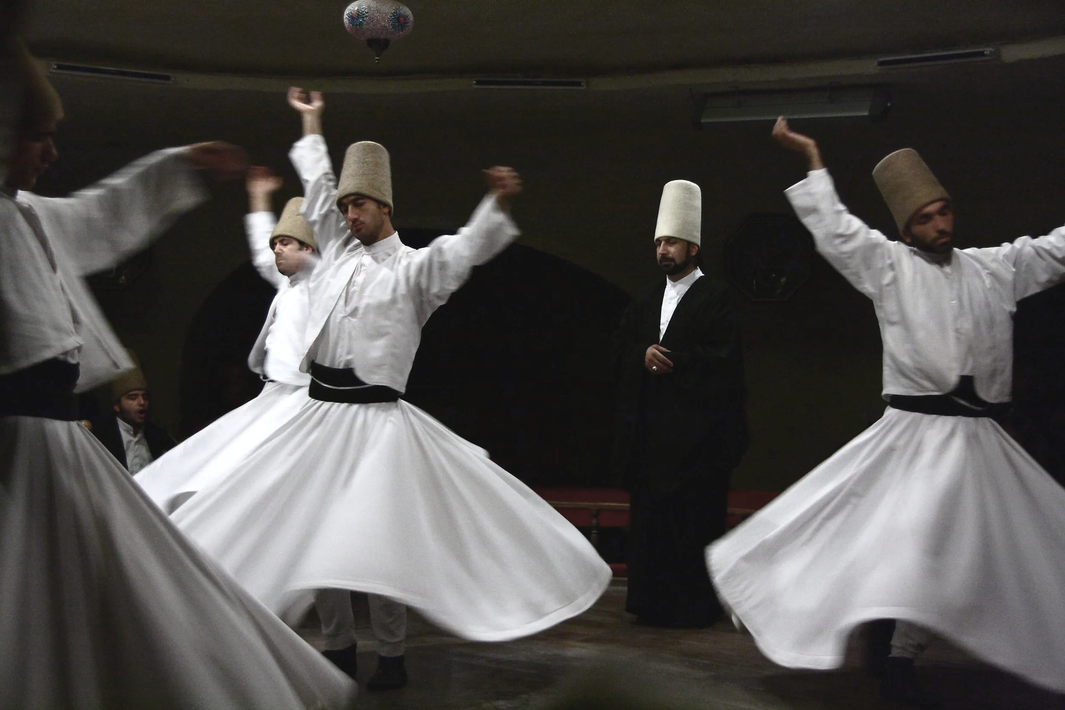 Men dressed in traditional white garments and tall hats are performing a whirling dance in a dimly lit room.